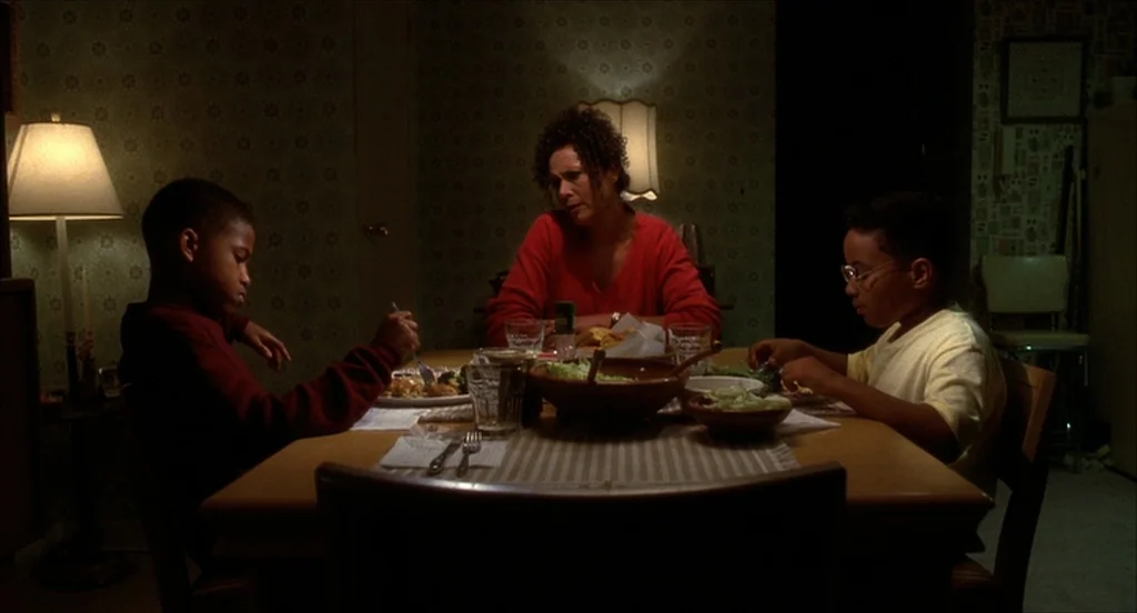 He Got Game (1998) movie still: medium — A dimly lit dining room features patterned wallpaper and a wooden table with food. A woman sits between two…