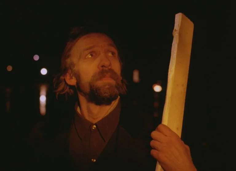 Russian Symphony (1994) movie still: medium — A man stands in extreme darkness with soft warm light illuminating his face. He holds a long piece of…