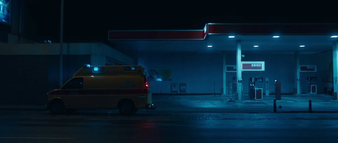 Broken Circle Breakdown (2012) movie still: wide — A quiet Esso gas station illuminated at night with a yellow ambulance parked nearby. The dark…