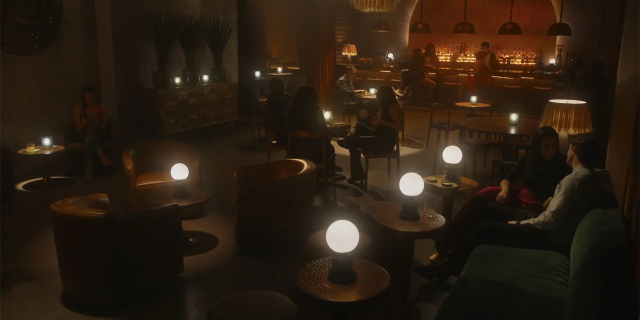 The Pickup (2025) movie still: wide — A dimly lit lounge with brown leather chairs and small tables topped with spherical lamps. Small groups of…