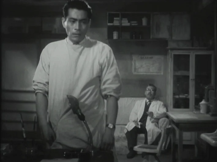 The Quiet Duel (1949) movie still: medium — A dimly lit room with medical equipment and a small cabinet. A man stands in the foreground as another…