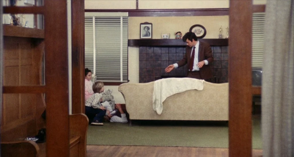 A Woman Under the Influence (1974) movie still: wide — A living room with dark wood trim, beige walls, and a tiled fireplace mantle. A man in a suit…