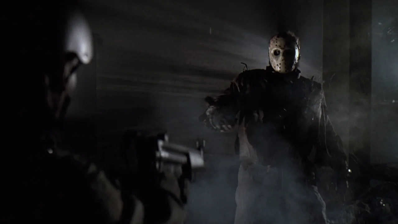 Jason X (2001) movie still: medium — A dimly lit indoor corridor filled with haze and deep shadows obscures the background. A large masked man…