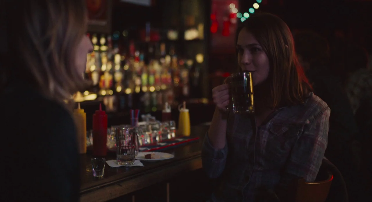 Mistress America (2015) movie still: medium — A dimly lit bar counter with rows of bottles and condiment dispensers in background. A woman sits at…
