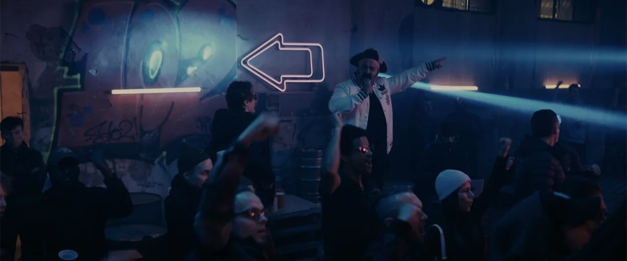 Perfect Addiction (2023) movie still: medium — A dimly lit industrial interior features graffiti walls and neon signage. A performer sings into a…