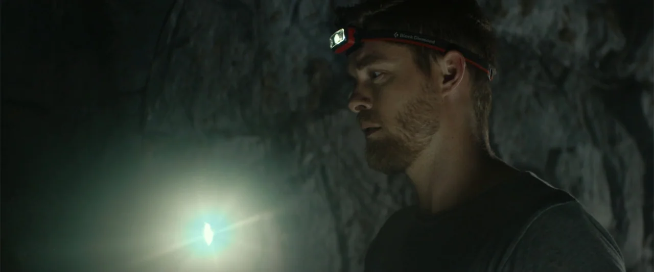Black Water: Abyss (2020) movie still: medium — A man stands within a dimly lit, damp rocky environment with uneven stone walls. He wears an active…