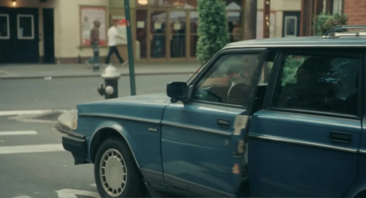 Motherhood (2020) movie still: medium — An urban intersection with a blue station wagon parked near a sidewalk. The vehicle has its front…
