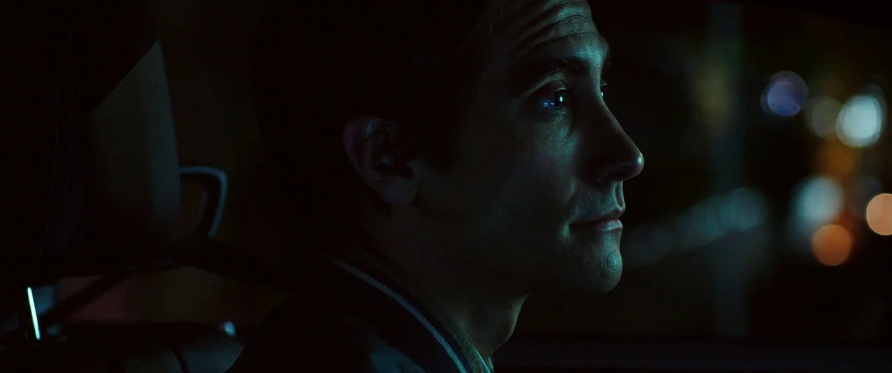 Nightcrawler (2014) movie still: closeup — A man sits inside a dark car at night. Faint city lights blur in the background through the window.