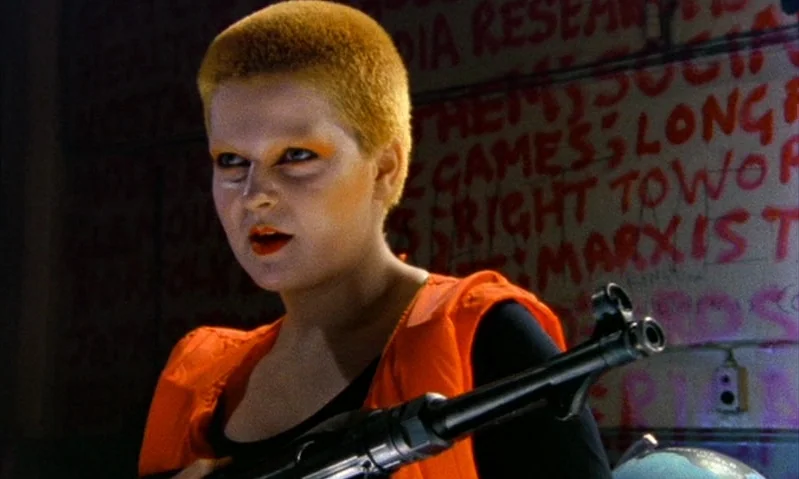 Jubilee (1978) movie still: medium — A dark indoor space features walls covered in hand-painted red graffiti lettering. A woman with short shaved…