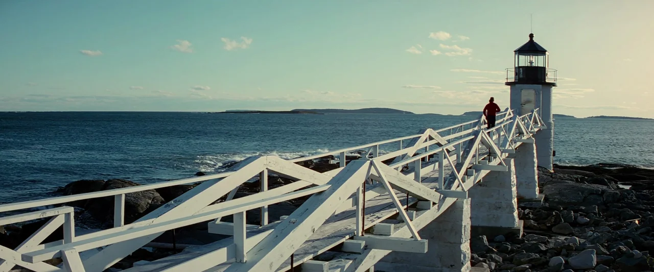Forrest Gump (1994) movie still: wide — A white lighthouse stands on a rocky coastline connected by a white wooden footbridge. A person in a red…