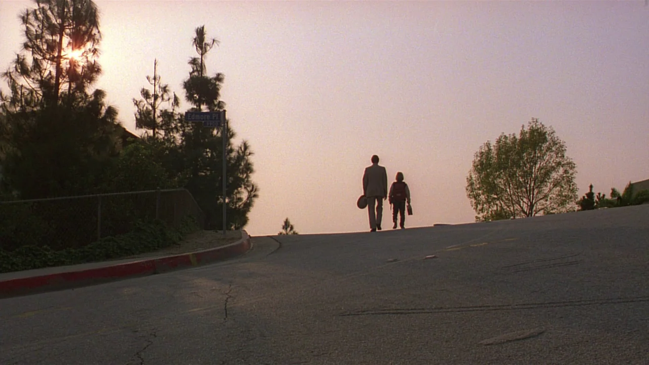 Paris, Texas (1984) movie still: wide — A residential street hill with asphalt, a street sign, and pine trees. An adult male and a young child walk…