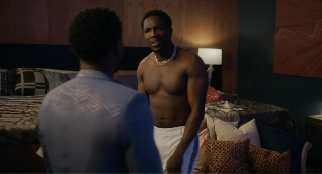 House Party (1990) movie still: medium — A hotel bedroom contains a large bed with pillows and wood paneling walls. One shirtless man stands facing…