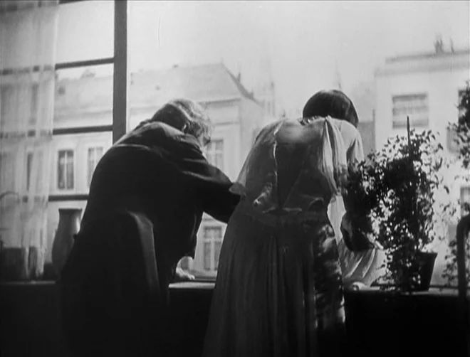 Pandora’s Box (1929) movie still: medium — A view from inside an apartment looking toward an urban residential street scene. A man and a woman stand…