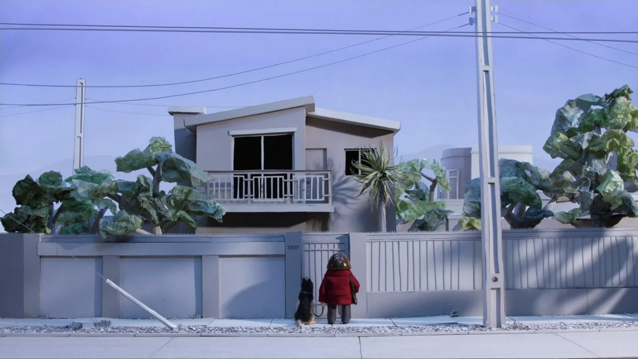 Bestia (2017) movie still: fullbody — A suburban house with a grey concrete fence stands behind a quiet paved sidewalk. A small person in a red coat…