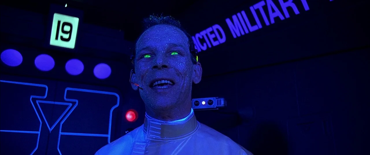 The Fifth Element (1997) movie still: medium — A dark, high-tech interior room features neon blue geometric outlines and glowing digital signage. A…