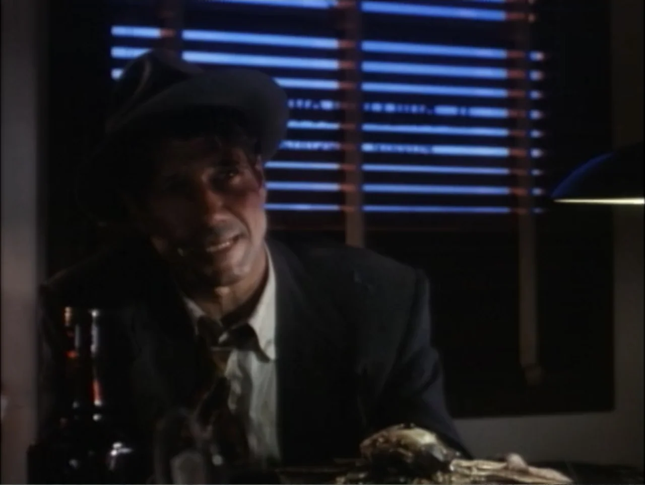 Cast a Deadly Spell (1991) movie still: medium — A dark office contains a desk with a bottle and blinds on the window. A man in a suit and hat sits…