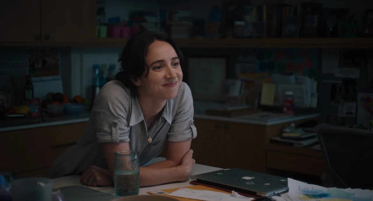 She Said (2022) movie still: medium — A dimly lit office space featuring wooden shelving filled with books and personal items. A woman rests her…
