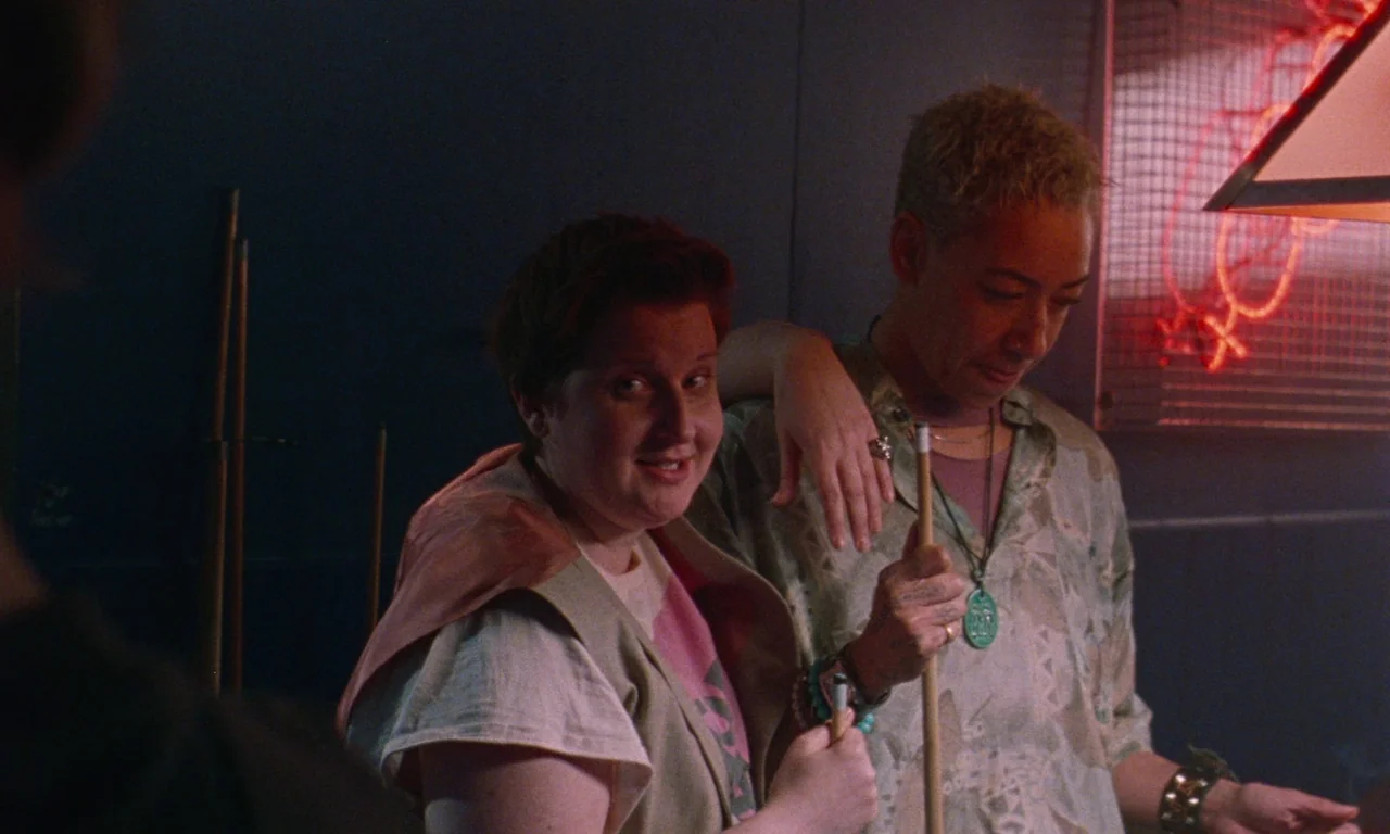 Blue Jean (2022) movie still: medium — A dimly lit room with red neon signs reflected on a dark wall. Two individuals stand together holding pool…