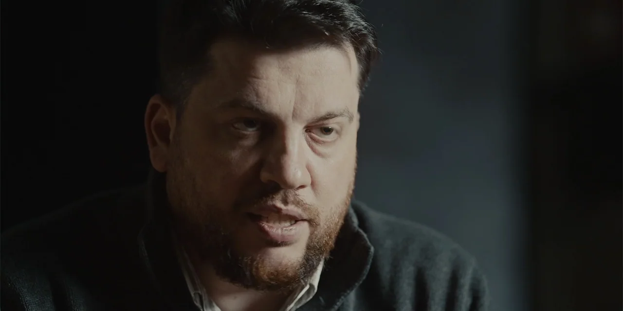 Navalny (2022) movie still: closeup — A man with a beard sits in a dimly lit room against a dark wall. He is mid-sentence, looking toward the camera…