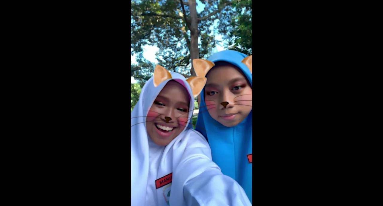 Tiger Stripes (2023) movie still: medium — Two teenage girls wearing headscarves stand outdoors with blurred green trees in the background. They…