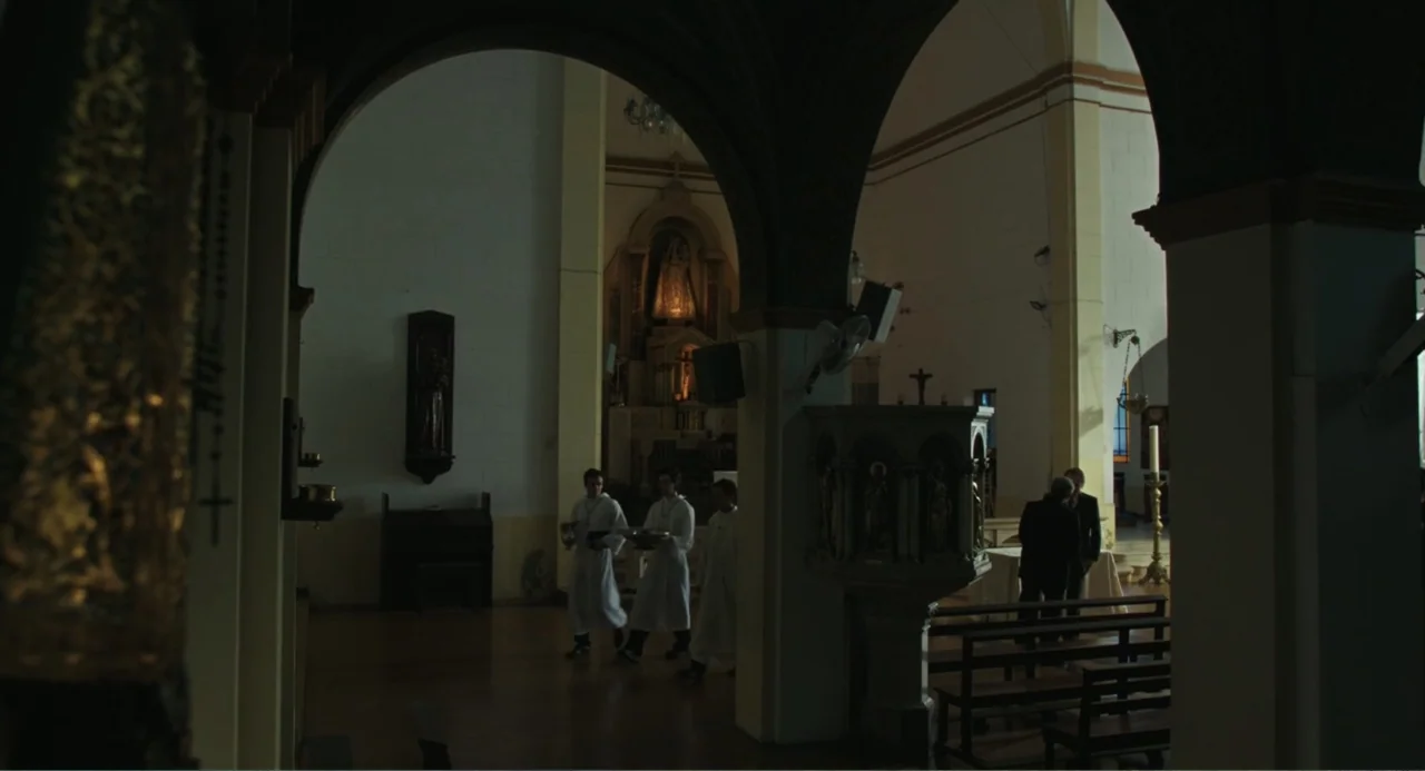 Belén (2025) movie still: wide — A spacious church interior features stone arches and wooden pews in dim light. Three figures dressed in white…