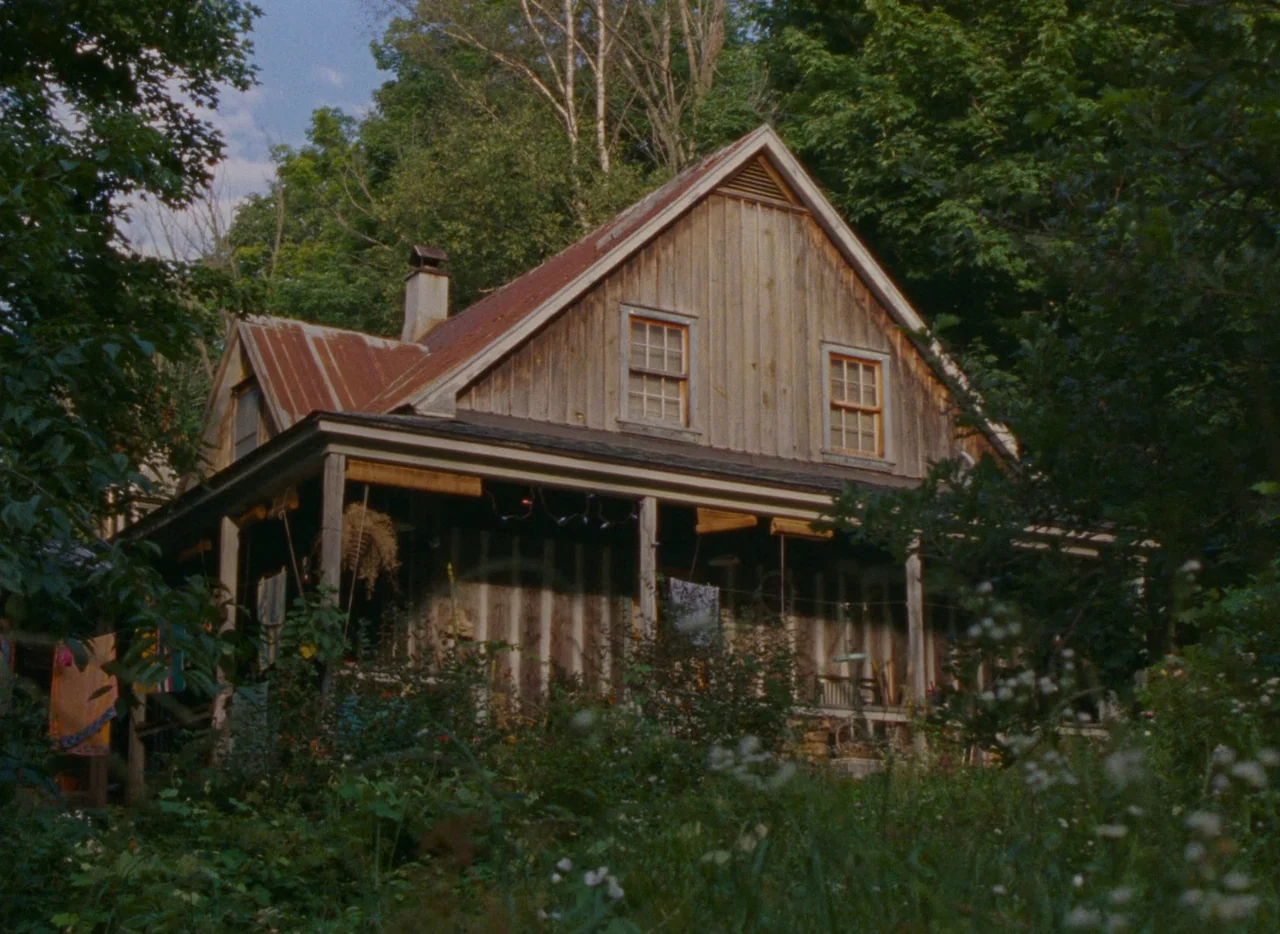 Falcon Lake (2022) movie still: establishing — A rustic wooden cottage with a weathered metal roof sits nestled in a forest. The house has a front…