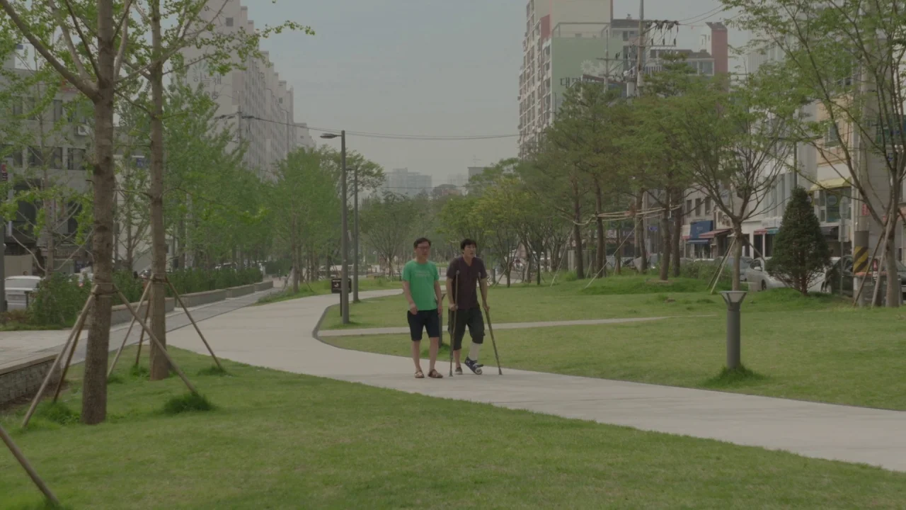 Yourself and Yours (2016) movie still: wide — A public park path winds through a residential neighborhood with apartment blocks in distance. Two men…