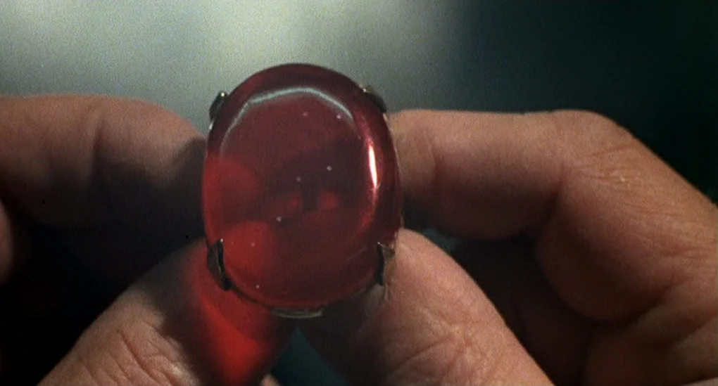 Blood From The Mummy’s Tomb (1971) movie still: closeup — A close-up of a silver ring featuring a large, oval-shaped translucent red stone. The ring…