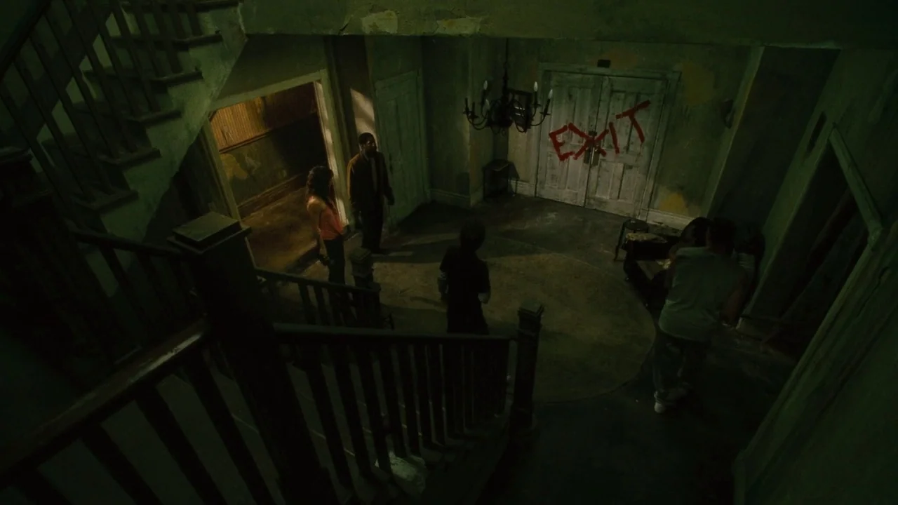 Saw II movie still: wide — A dilapidated Victorian foyer features peeling paint, a wooden staircase, and graffiti on doors. Four individuals stand…