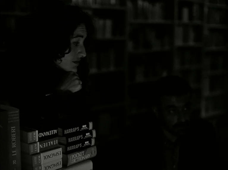 Eloge de L’amour (2001) movie still: medium — A dark, dimly lit library with shelves of books visible in the blurred background. A woman in the…
