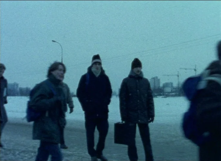From The East (1993) movie still: fullbody — A snowy, grey urban landscape features distant construction cranes against an overcast sky. Three young…