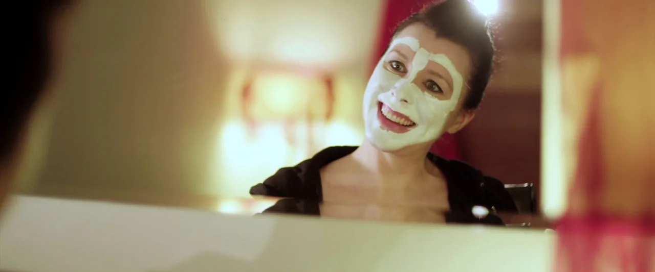 Prevenge (2016) movie still: closeup — A woman stands before a mirror in a brightly lit space. She smiles at her reflection while wearing a thick…