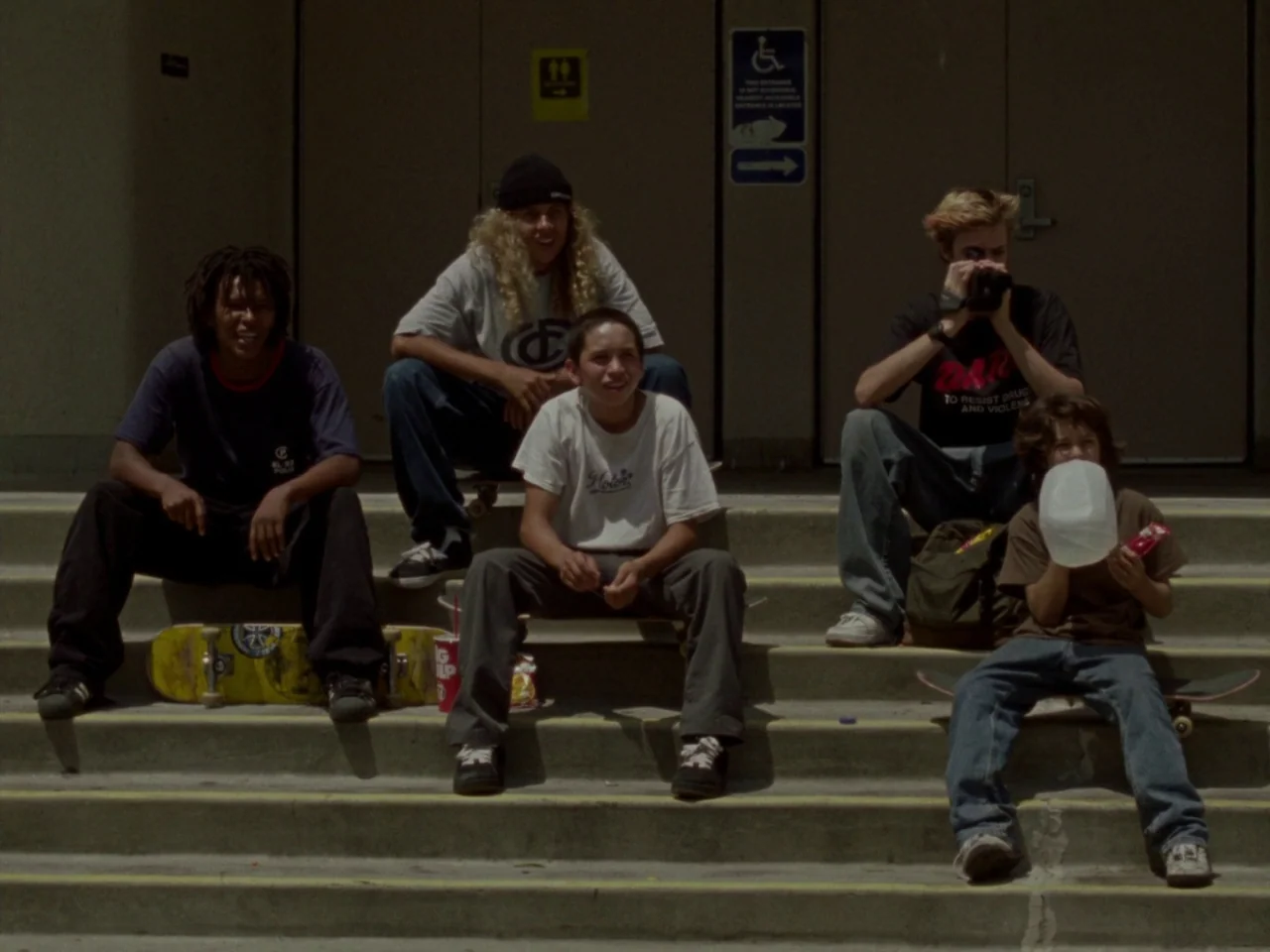 Mid90s (2018) movie still: fullbody — Five teenagers and a young child sit together on a concrete staircase outdoors. They are accompanied by…