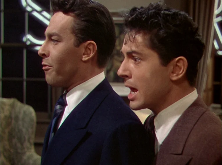 Rope (1948) movie still: medium — A furnished interior room has a dark window frame with a neon sign outside. Two men stand close together in side…