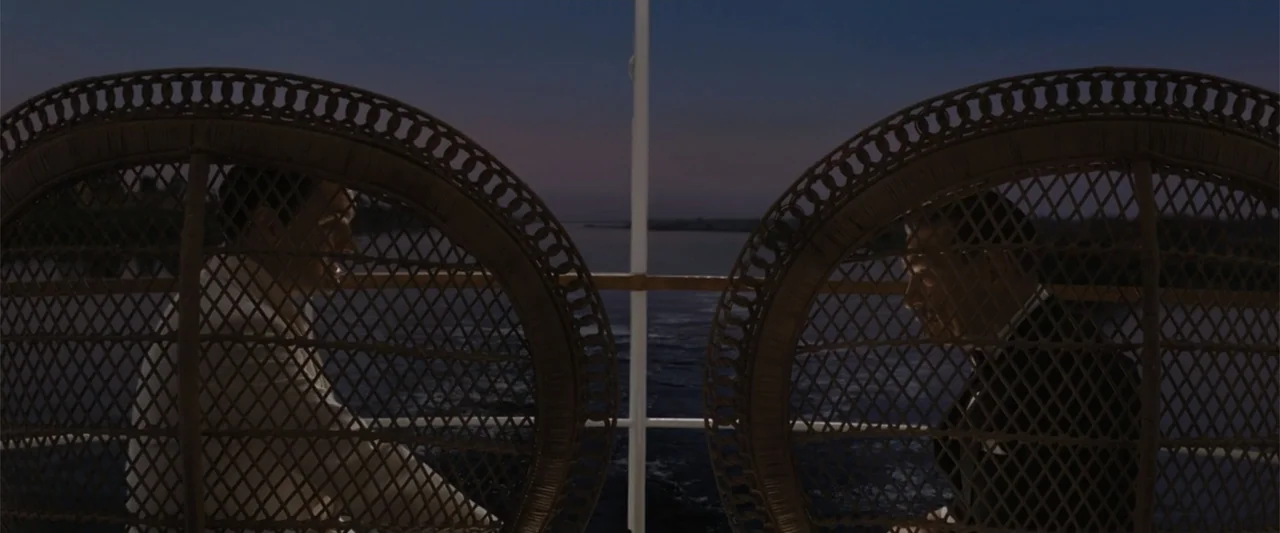 Death on the Nile (2022) movie still: medium — Two men sit in large wicker chairs on the back deck of a boat. The Nile River flows behind them under…