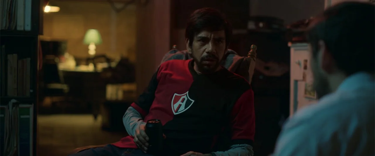 Tequila Re-Pasado (2023) movie still: medium — A dimly lit room contains a man seated in a wooden chair while another person is seen from behind.…