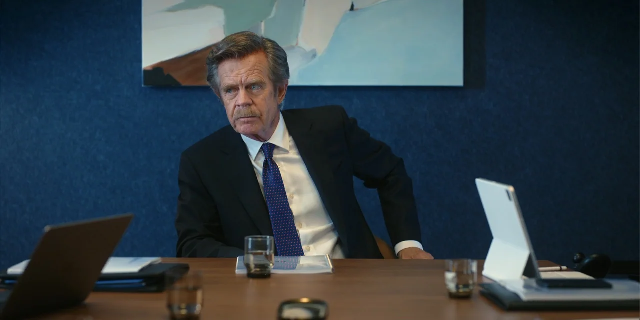 Ricky Stanicky (2024) movie still: medium — A dark blue wall serves as the background for a wood-topped conference table. An older man in a suit…