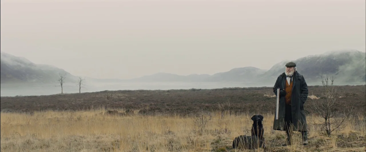 Skyfall (2012) movie still: wide — A vast, flat, overcast moorland stretches toward misty mountains in the distance. An elderly man stands in the…