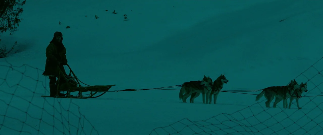 Siberia (2019) movie still: wide — A snowy landscape stretches out under a dark, uniform teal-tinted moonlight. A man stands on a wooden sled behind…