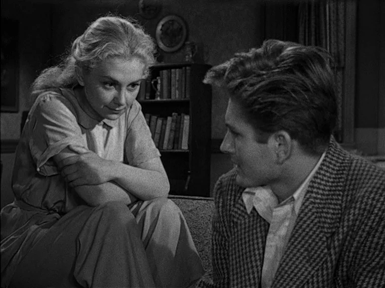The Big Night (1951) movie still: medium — A dimly lit room features a wooden bookshelf filled with books behind a sofa. A woman in a dress and a…