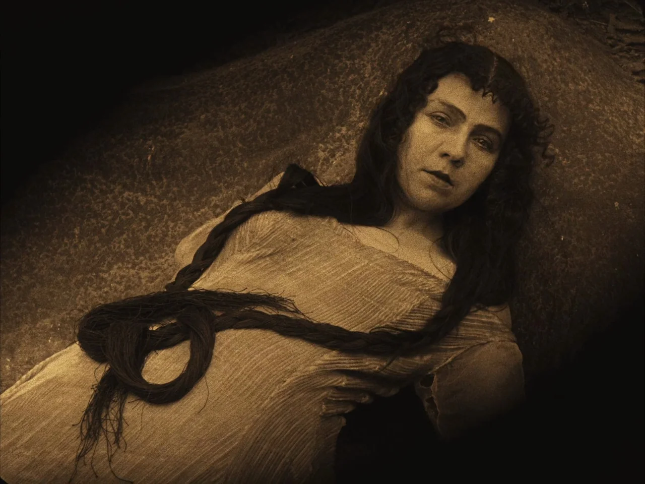 The Golem (1920) movie still: medium — A woman lies on a textured, dark rock surface in an outdoor setting. She stares forward while her long dark…