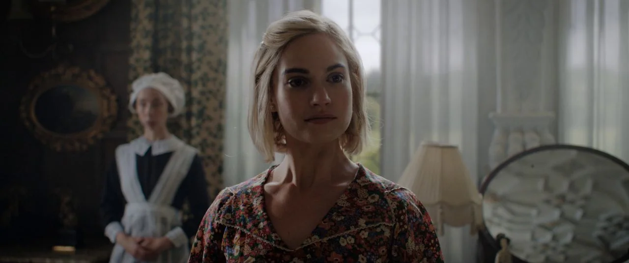 Rebecca movie still: medium — A furnished interior space with a window, a vanity mirror, and a table lamp. A woman in a floral blouse stands in…