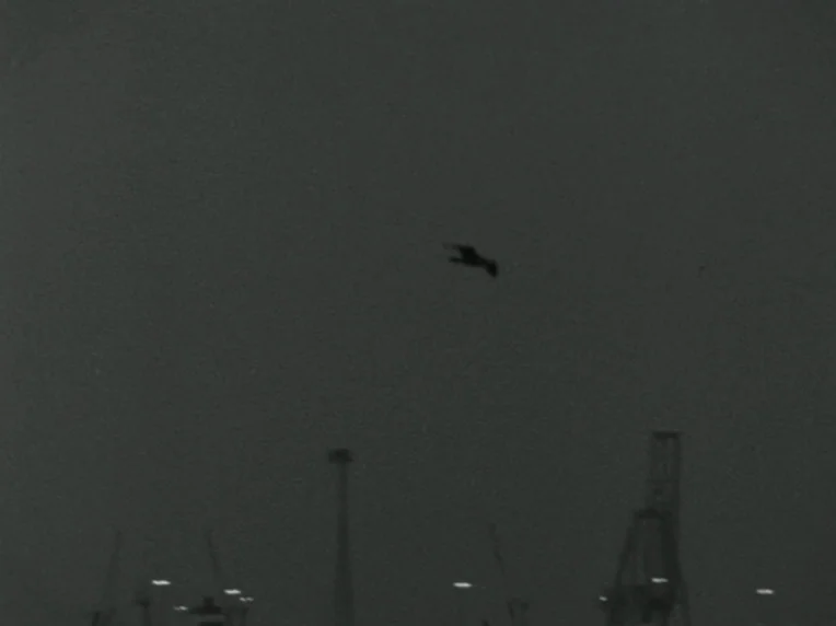 Madonna and Child (1980) movie still: wide — A dark, overcast sky hangs above an industrial port at night. A lone bird flies across the center of…