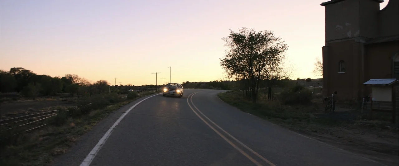 Trap House (2025) movie still: wide — A rural paved road curves past an old adobe building at the side of a landscape. A car drives towards the…