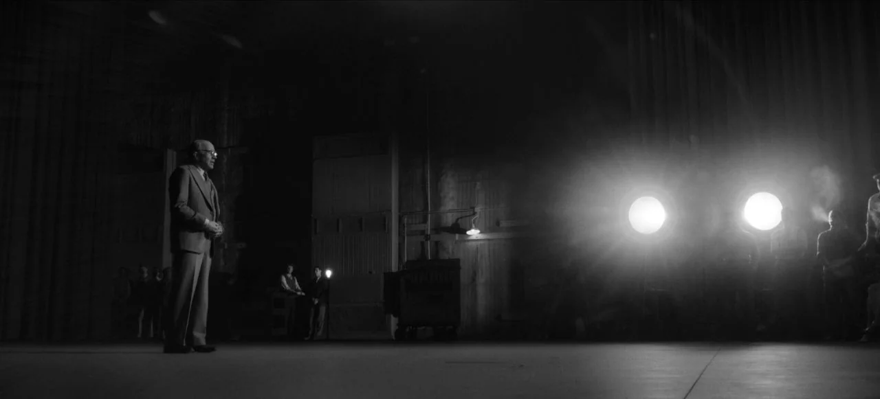 Mank (2020) movie still: wide — A spacious film studio interior with industrial equipment and two bright spotlights burning. An elderly man in a…
