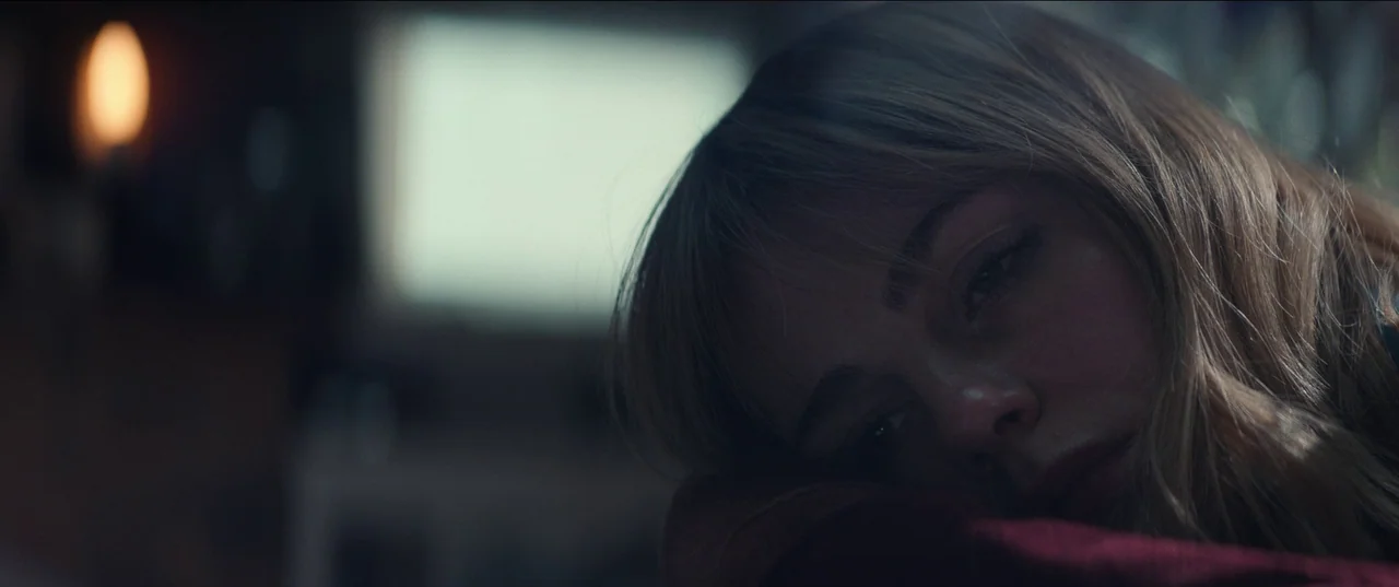 The Greatest Hits (2024) movie still: closeup — A dimly lit indoor space with blurred furniture in the background. A young woman rests her cheek…