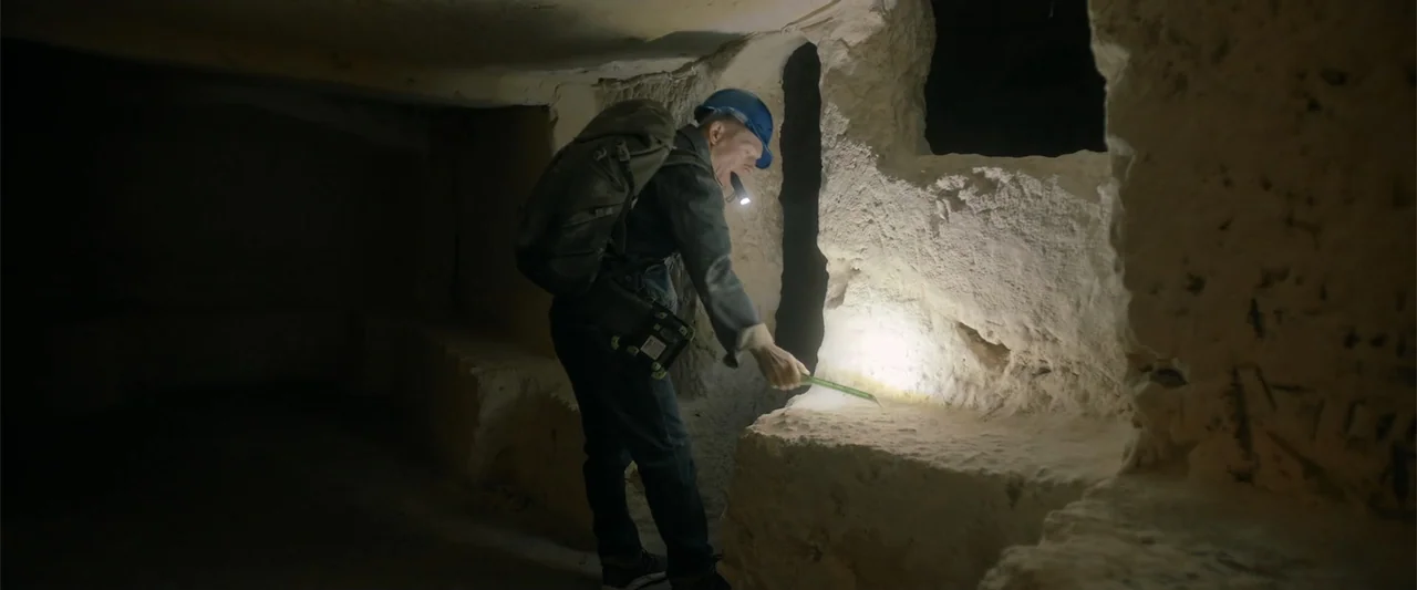 Hounds of War (2024) movie still: medium — A narrow limestone passage with rough, unrefined walls and a dark ceiling. A man in work gear uses a…
