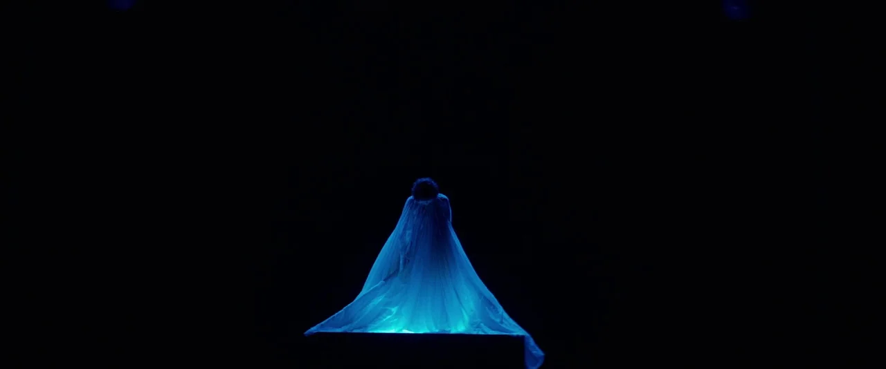 The Dancer movie still: wide — A dark, empty theatrical stage houses a lone figure shrouded in light. The subject stands on a platform with a long…