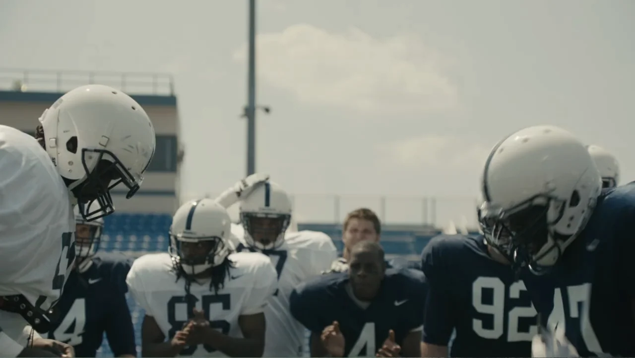 Paterno (2018) movie still: medium — A football field with blue stadium bleachers visible in the background under bright daylight. Several football…