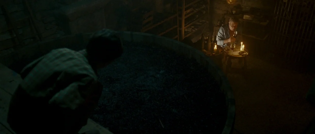 A Good Year (2006) movie still: medium — A dimly lit cellar contains a large wooden vat filled with dark grapes. One man sits at a small table lit…